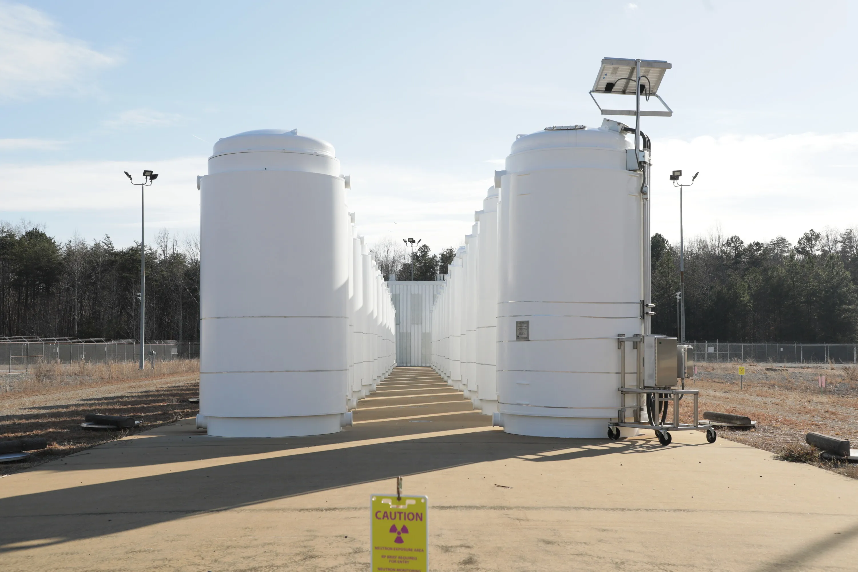 Two rows of steel and concrete dry storage casks line a fenced concrete pad, the standard way used nuclear fuel is stored once it leaves the plant's cooling pool.