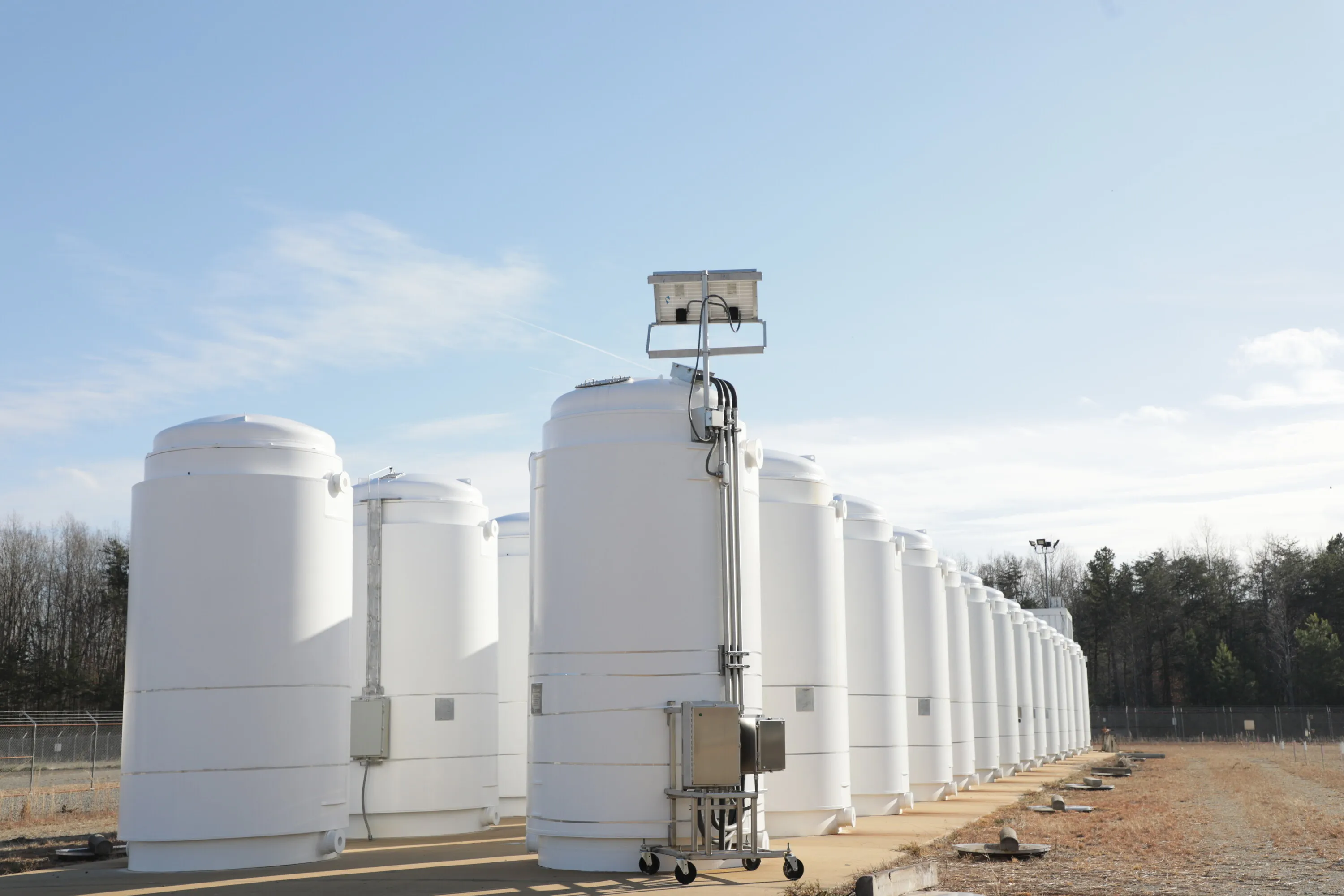 A row of sealed dry storage casks holding used nuclear fuel, led by an instrumented research cask that records how high burnup fuel behaves over years in storage.