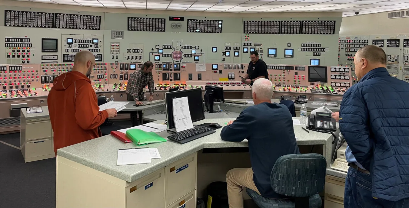 Licensed operators work at the main control boards in the Palisades control room while a supervisor reviews procedures at the desk.
