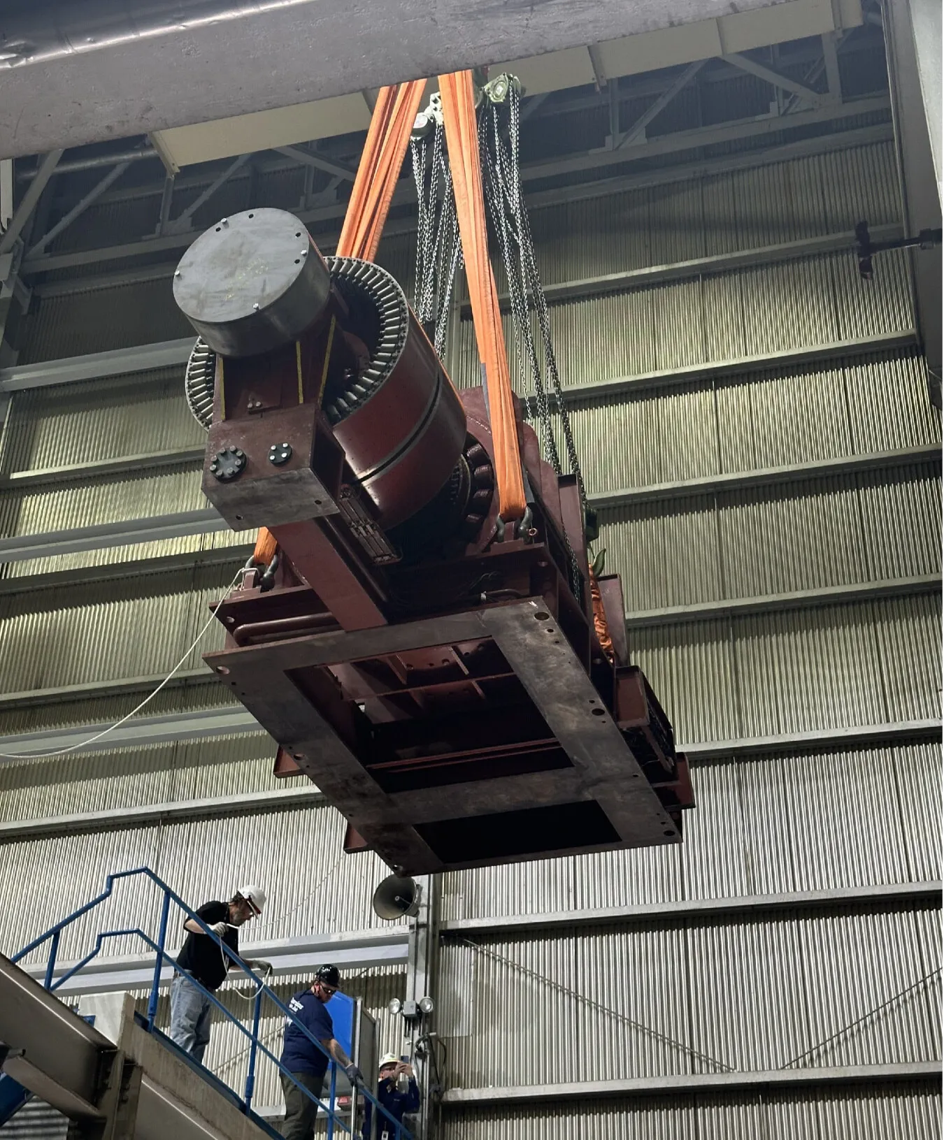 An overhead crane lifts a large motor assembly inside Palisades Nuclear Plant as workers guide it from a stairway during work to return the plant to service.