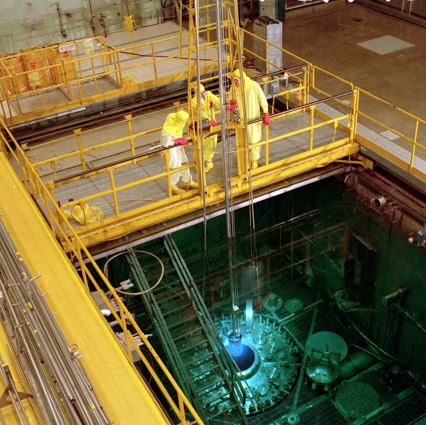 Workers in protective clothing stand on a refueling bridge above a reactor pool, where several meters of water shield them while long-handled tools reach the core below.